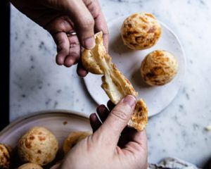 A close-up of several Pão de Queijo on a baking sheet. The golden rolls have a slight crackle on their surface and are scattered with a light dusting of tapioca flour.