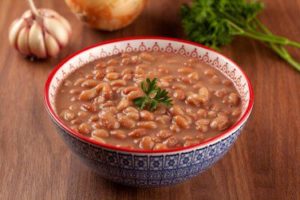 A close-up view of a steaming bowl filled with tender brown beans nestled in a savory broth. Chopped fresh herbs garnish the top, with a side of fluffy white rice in the background.