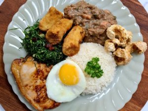 A vibrant plate showcasing Virado à Paulista – creamy tutu bean stew nestled next to juicy pork steaks, sausages, fried bananas, and crispy pork rinds.